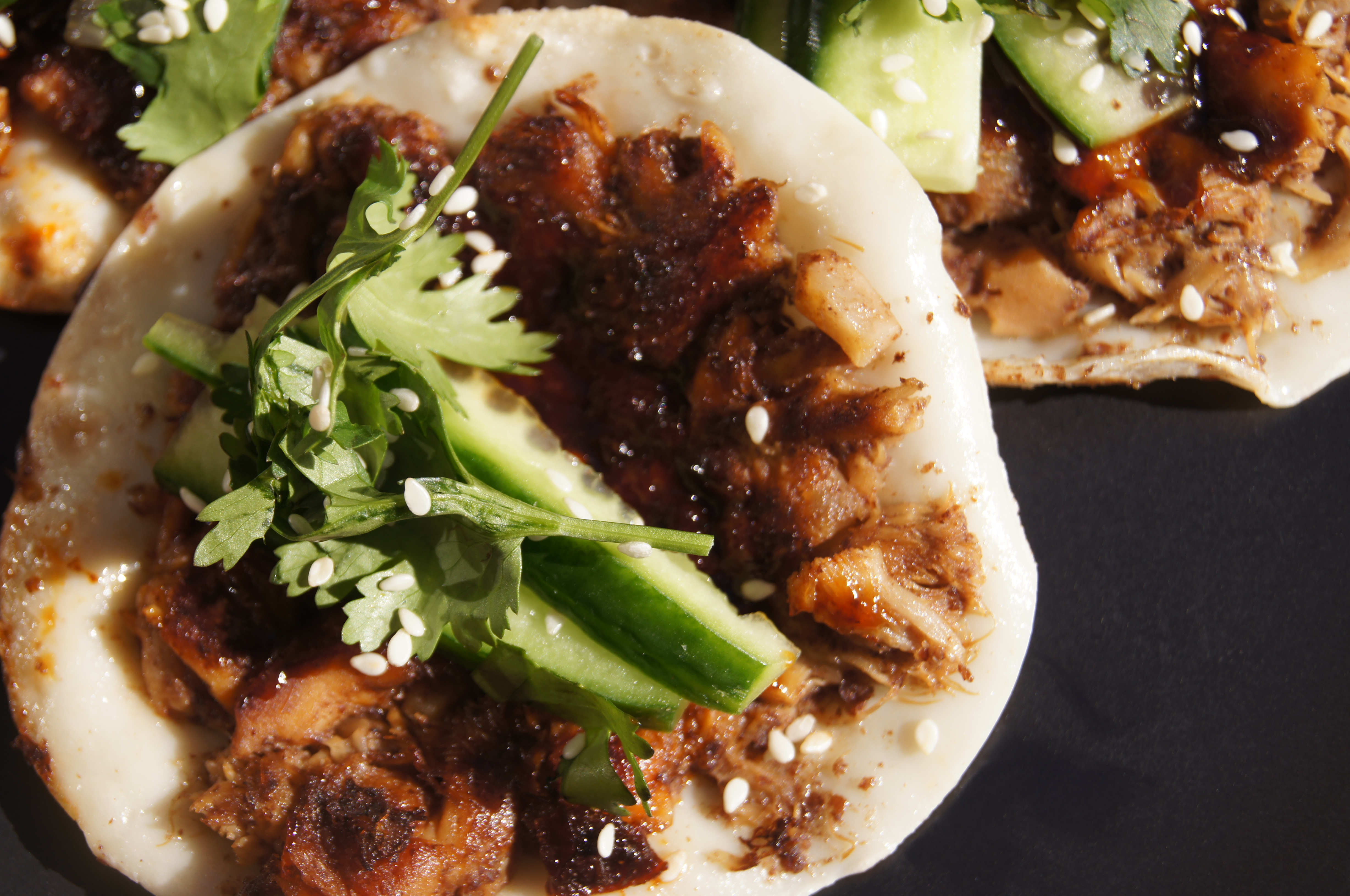 Close-up of crispy dumplings topped with fresh cucumber slices, coriander, and sesame seeds on a black plate.
